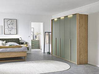 Malin: Vorschlag 701, Balkeneiche furniert, Absetzung Lack Jungle Green, bestehend aus Drehtürenschrank 6-türig, 3 Griffstangen in Eiche massiv, ca. B 299, H 229, T 60 cm, Doppelbett in Luxushöhe, ca. B 189, H 105, T 222 cm, Liegefläche ca. 180 x 200 cm, BSH 48,6 cm, Polsterkopfteil "Luna" in Stoff Maranta Jungle Green, 100 % Polyester, Fußteil mit Lisene, unten abgerundet und Kufe in Eiche, 2 Hängekonsolen mit je 1 Schubkasten, jeweils ca. B 60, H 23, T 46 cm. Zubehör: Passepartout ohne Beleuchtung (903004); 3er-Set LED-Stabbeleuchtung (90353); Paneelaufsatz links und rechts mit Holzboden (20216, 20226); indirekte LED-Holzbodenbeleuchtung (80027); 2er-Set LED-Flex-Leuchte Anthrazit, USB-Anschluss (80033). 